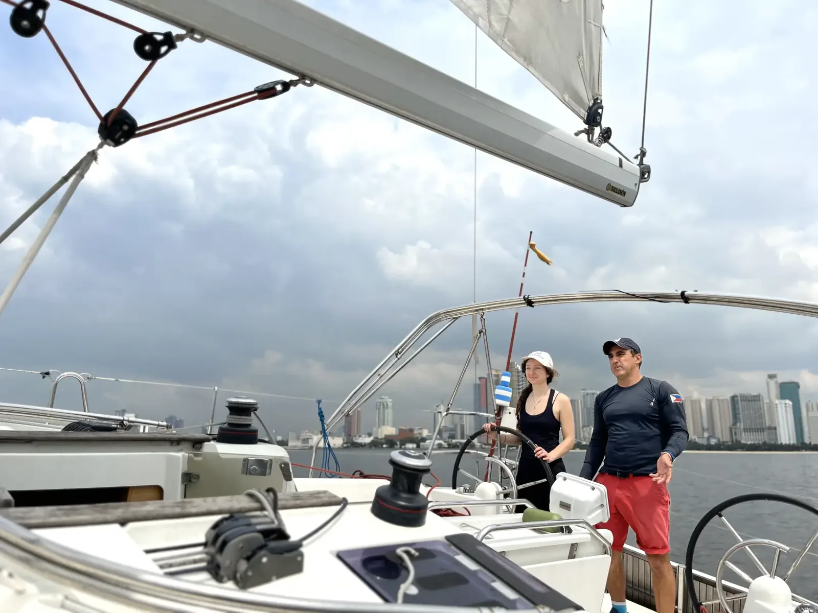 Two sailors standing in the cockpit of a yacht under sail with a city skyline in the distance